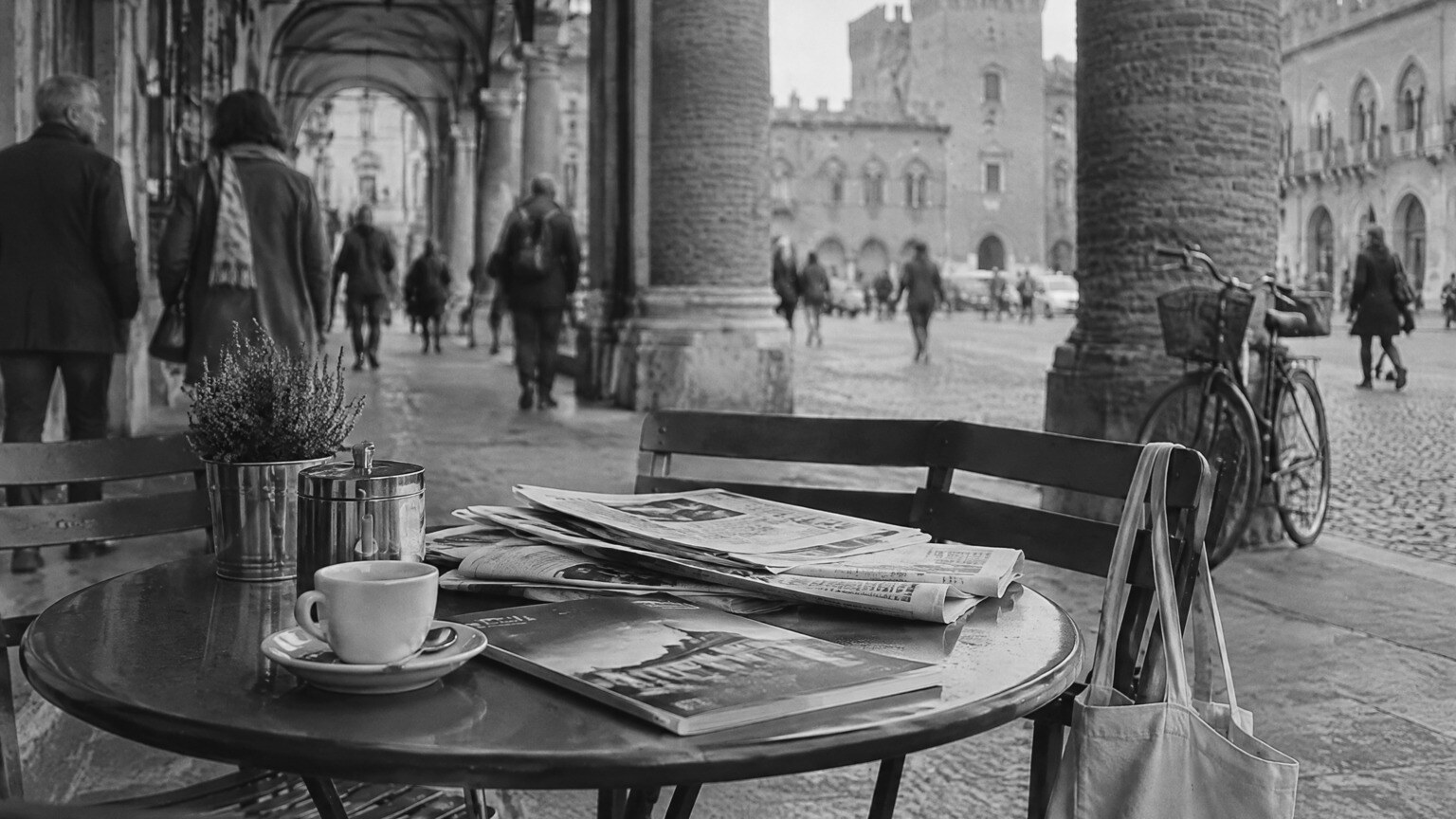 In bianco e nero, un tavolino di un caffè sotto i portici di Ferrara con giornali piegati, una rivista e una tazzina; sullo sfondo passanti, una bicicletta e i palazzi del centro
